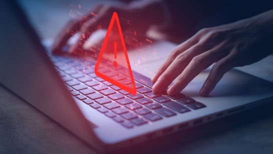 Hands typing on a laptop keyboard with a red warning symbol displayed on the screen