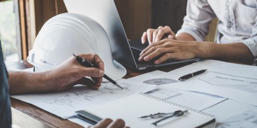 Engineers collaborating on blueprints with a laptop and safety helmet, representing information systems management for engineering firms in Central Ontario