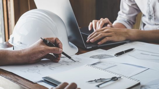Engineers collaborating on blueprints with a laptop and safety helmet, representing information systems management for engineering firms in Central Ontario