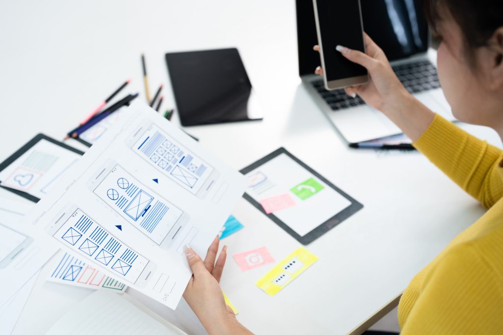 Designer reviewing mobile app wireframes while holding a smartphone at a workspace