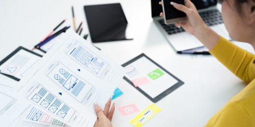 Designer reviewing mobile app wireframes while holding a smartphone at a workspace