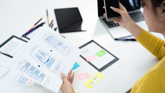 Designer reviewing mobile app wireframes while holding a smartphone at a workspace