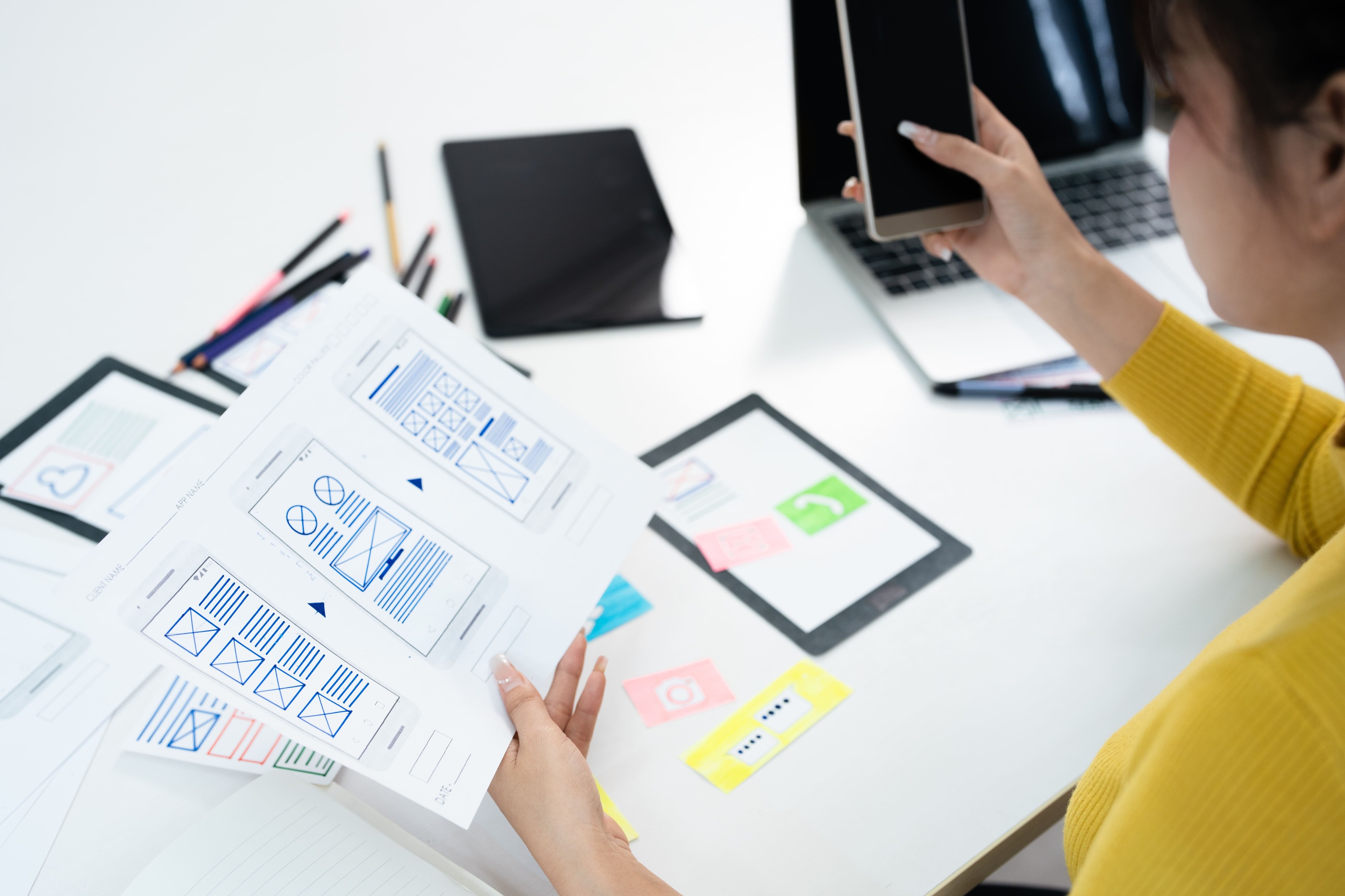 Designer reviewing mobile app wireframes while holding a smartphone at a workspace