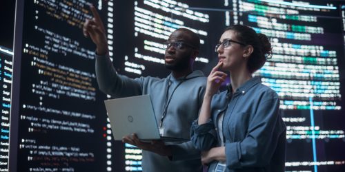 Two programmers analyzing code on a large screen, one pointing while holding a laptop
