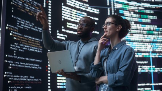 Two programmers analyzing code on a large screen, one pointing while holding a laptop