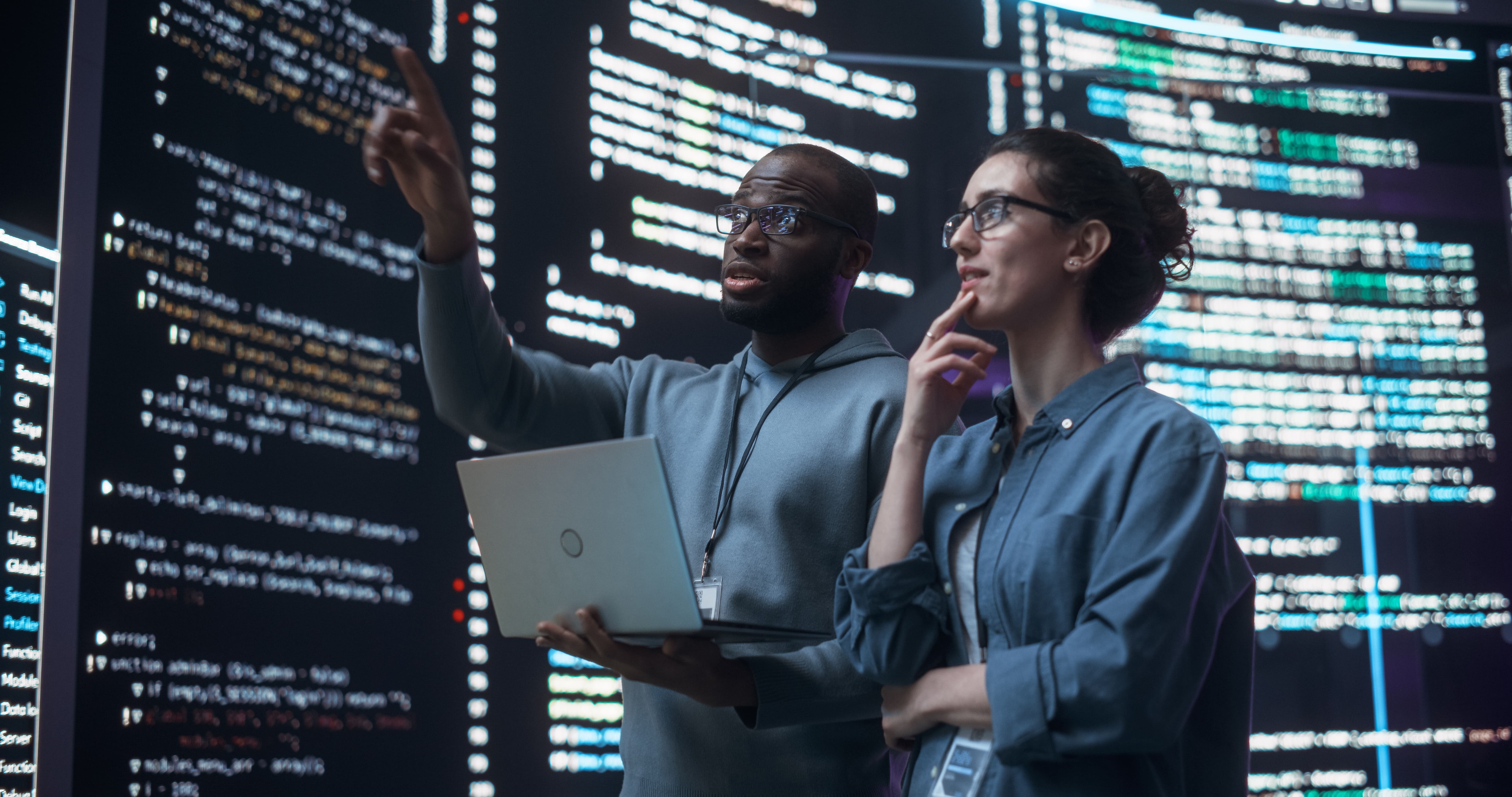 Two programmers analyzing code on a large screen, one pointing while holding a laptop