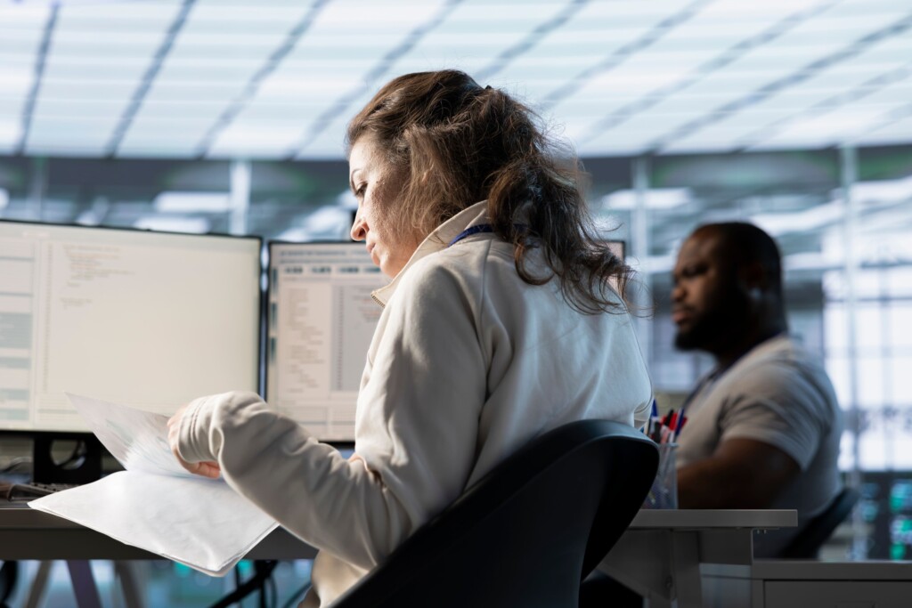 Woman reviewing documents at a desk with computer screens, while a colleague works in the background.