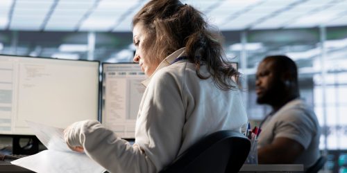 Woman reviewing documents at a desk with computer screens, while a colleague works in the background.