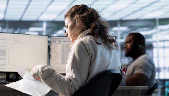 Woman reviewing documents at a desk with computer screens, while a colleague works in the background.