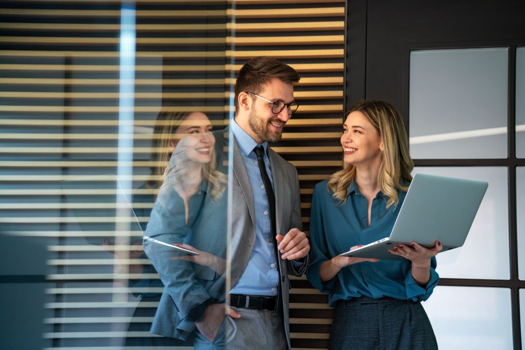 Two colleagues smiling and discussing work while standing in an office; the woman holds an open laptop, and the man, dressed in a suit, listens attentively beside a glass wall with blinds.