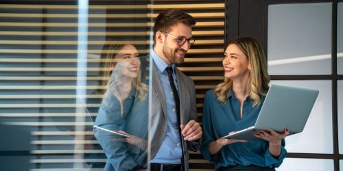 Two colleagues smiling and discussing work while standing in an office; the woman holds an open laptop, and the man, dressed in a suit, listens attentively beside a glass wall with blinds.