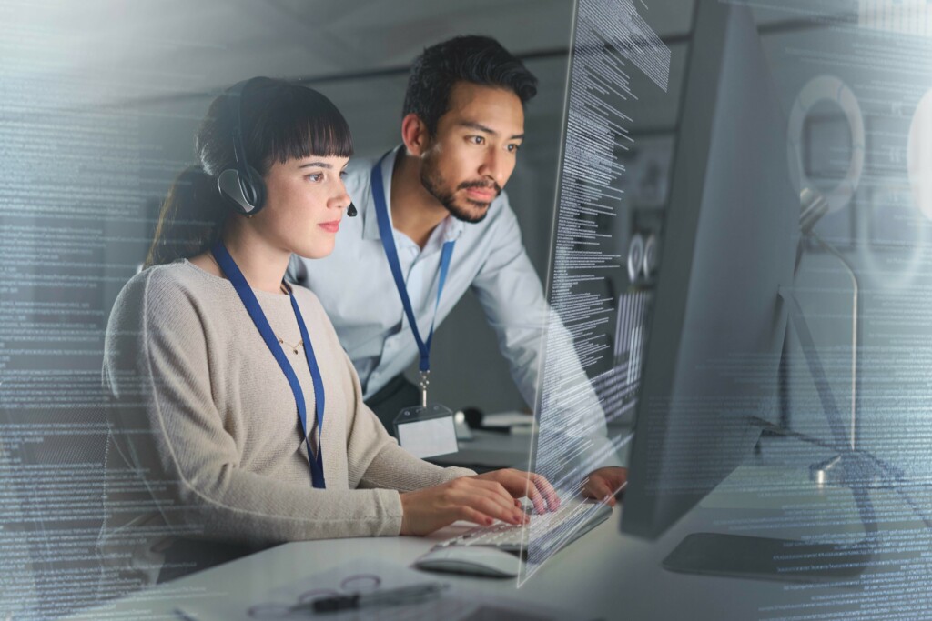Two IT professionals working together at a computer workstation, analyzing code displayed on a large transparent screen; the woman types while wearing a headset, and the man observes closely beside her.