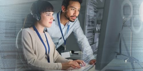 Two IT professionals working together at a computer workstation, analyzing code displayed on a large transparent screen; the woman types while wearing a headset, and the man observes closely beside her.