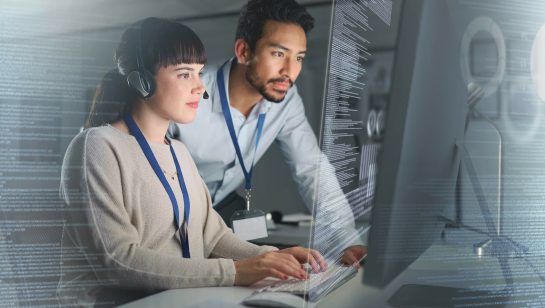 Two IT professionals working together at a computer workstation, analyzing code displayed on a large transparent screen; the woman types while wearing a headset, and the man observes closely beside her.