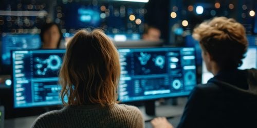 Two people working side by side in a dimly lit control room, looking at large computer monitors displaying complex data visualizations and system analytics.