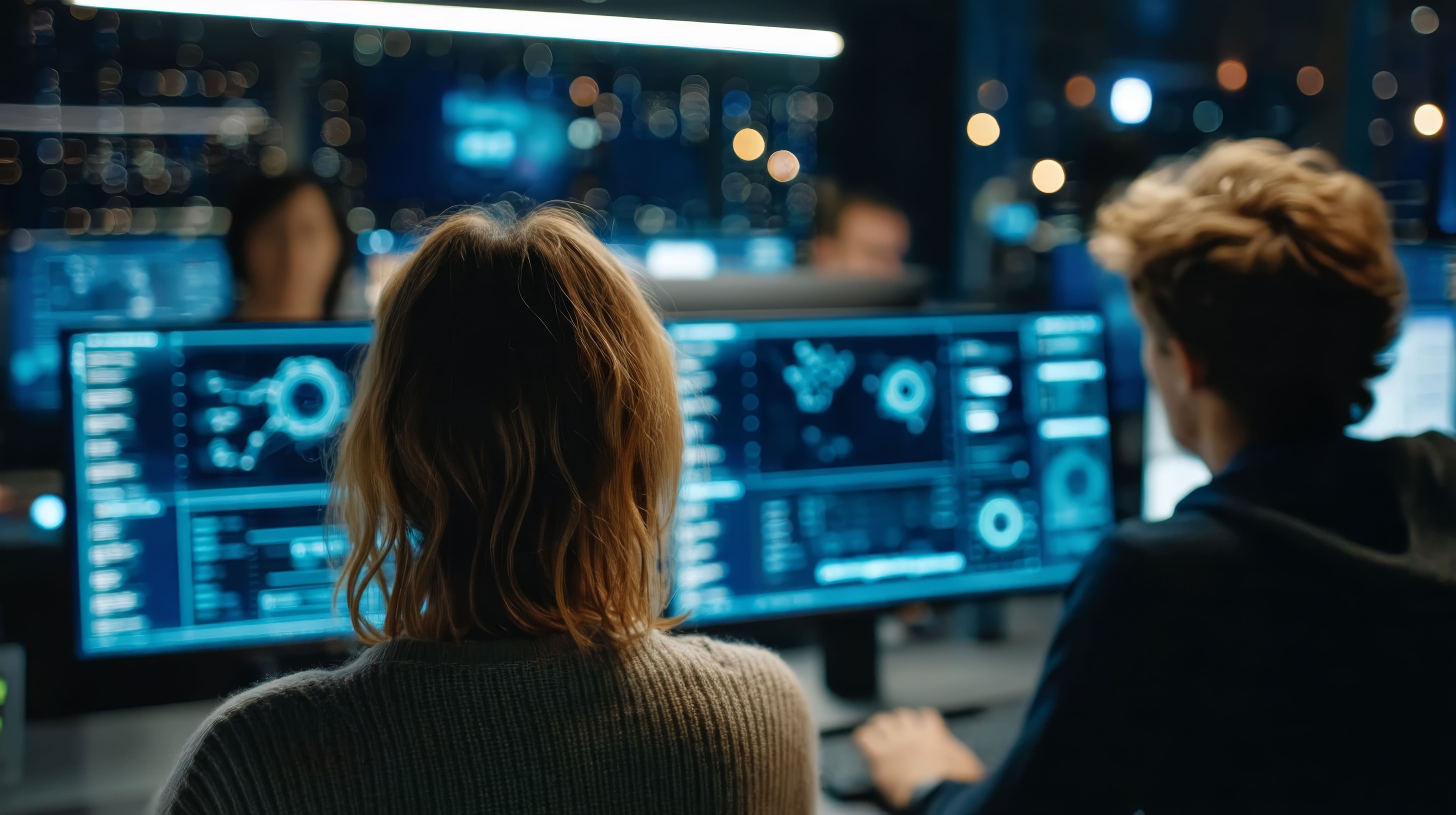 Two people working side by side in a dimly lit control room, looking at large computer monitors displaying complex data visualizations and system analytics.