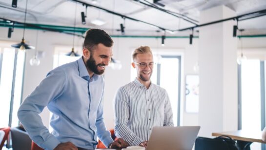Two smiling businessmen standing at a desk and looking at a laptop in a modern office.