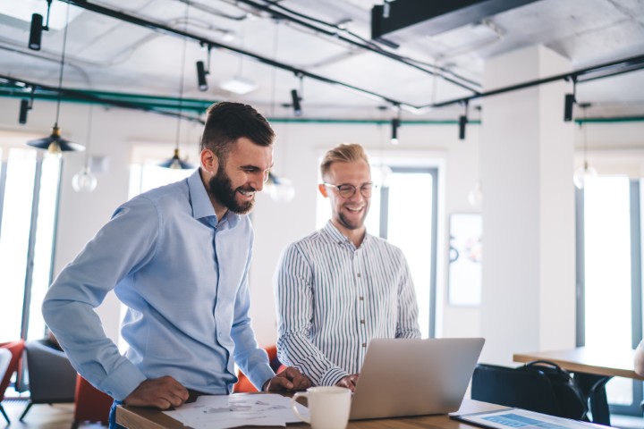Two smiling businessmen standing at a desk and looking at a laptop in a modern office.