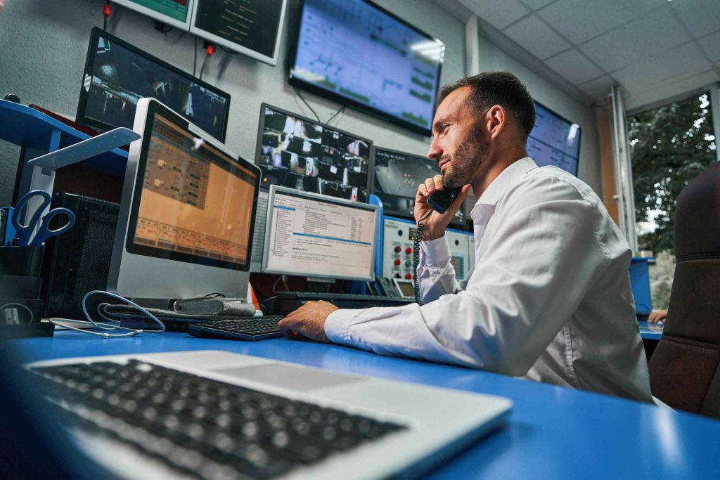 Man in a control room working on multiple computer monitors while talking on the phone
