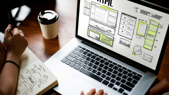 Laptop displaying HTML website wireframes on a desk with notes and a coffee cup, representing the web design process