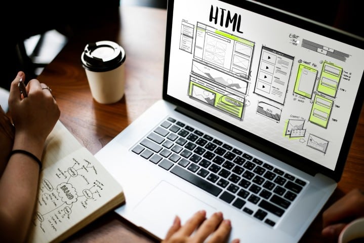 Laptop displaying HTML website wireframes on a desk with notes and a coffee cup, representing the web design process