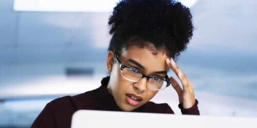 Professional woman wearing glasses looking stressed while working on a laptop in an office environment.