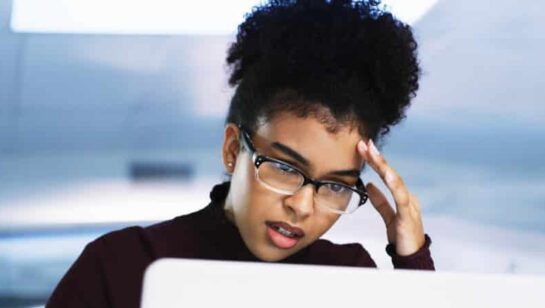 Professional woman wearing glasses looking stressed while working on a laptop in an office environment.