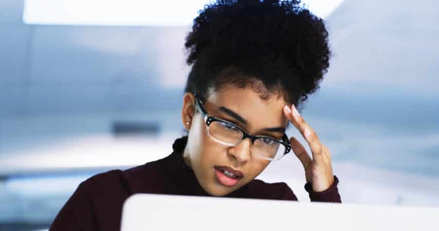 Professional woman wearing glasses looking stressed while working on a laptop in an office environment.