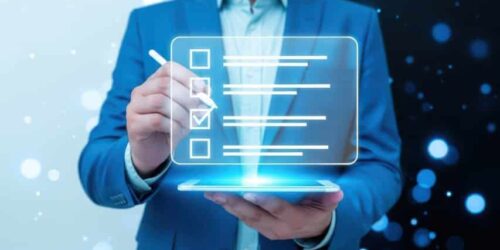 A person in a blue business suit holds a glowing digital tablet that projects a transparent, holographic checklist into the air. The person uses a stylus to mark a checkbox on the floating interface. The background features a split design with soft white bokeh lights on the left and sharp blue digital bokeh on the right, symbolizing the intersection of business and modern technology.
