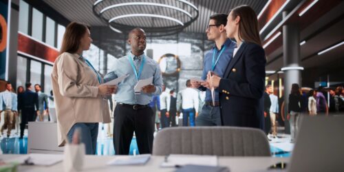 A diverse group of four professionals stand in the center of a bustling, modern office lobby discussing documents. The man in the light blue shirt is speaking while holding papers, while his colleagues listen attentively. They are all wearing blue lanyards around their necks. In the blurred background, other office workers move through the large, open space, which features high ceilings, circular light fixtures, and glass partitions. The foreground shows a desk with office supplies, including a tablet and a notebook, slightly out of focus.