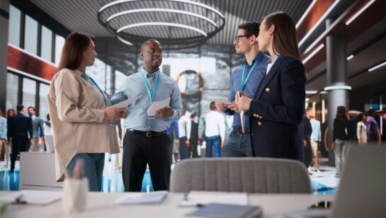 A diverse group of four professionals stand in the center of a bustling, modern office lobby discussing documents. The man in the light blue shirt is speaking while holding papers, while his colleagues listen attentively. They are all wearing blue lanyards around their necks. In the blurred background, other office workers move through the large, open space, which features high ceilings, circular light fixtures, and glass partitions. The foreground shows a desk with office supplies, including a tablet and a notebook, slightly out of focus.