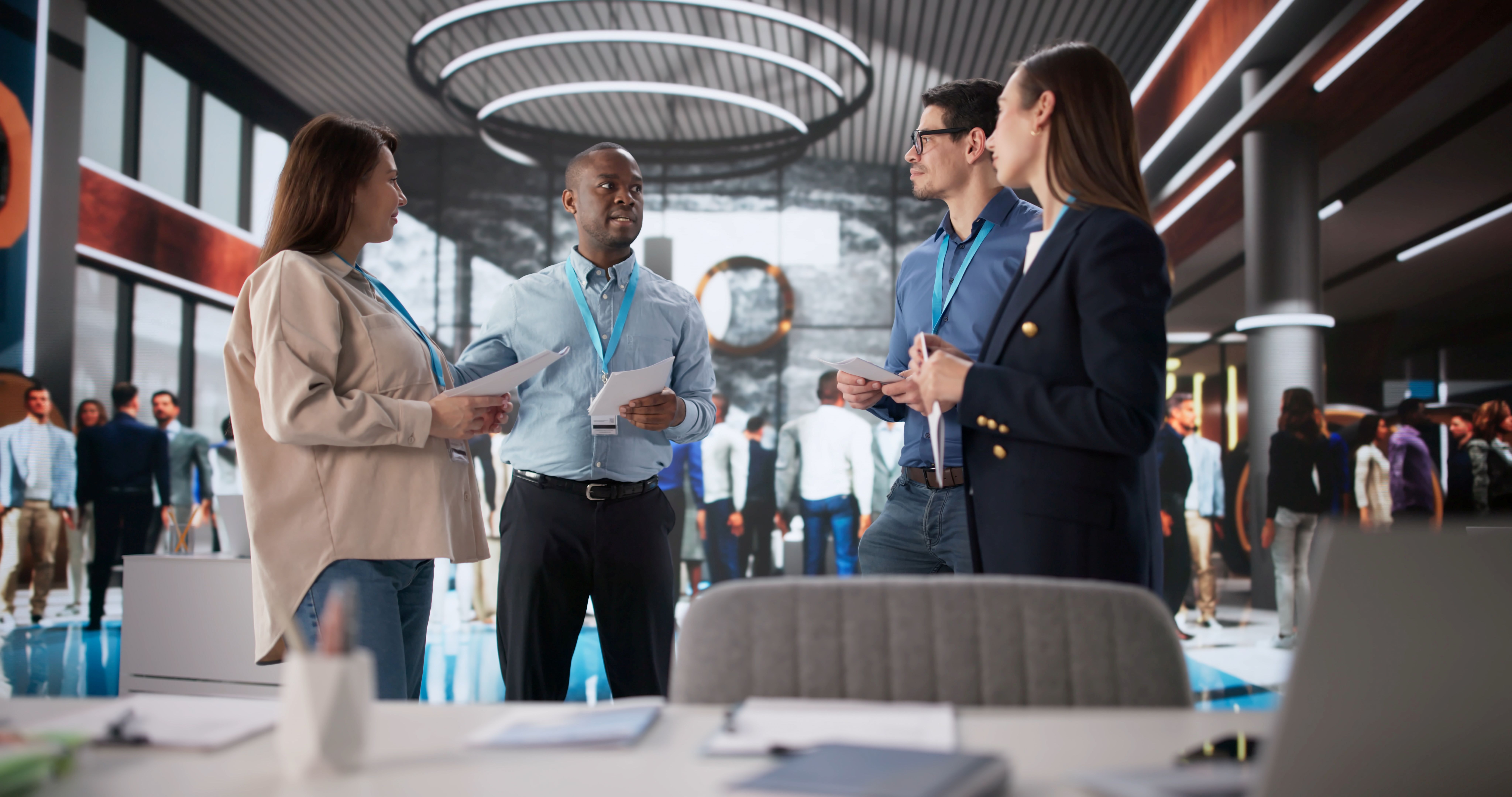 A diverse group of four professionals stand in the center of a bustling, modern office lobby discussing documents. The man in the light blue shirt is speaking while holding papers, while his colleagues listen attentively. They are all wearing blue lanyards around their necks. In the blurred background, other office workers move through the large, open space, which features high ceilings, circular light fixtures, and glass partitions. The foreground shows a desk with office supplies, including a tablet and a notebook, slightly out of focus.