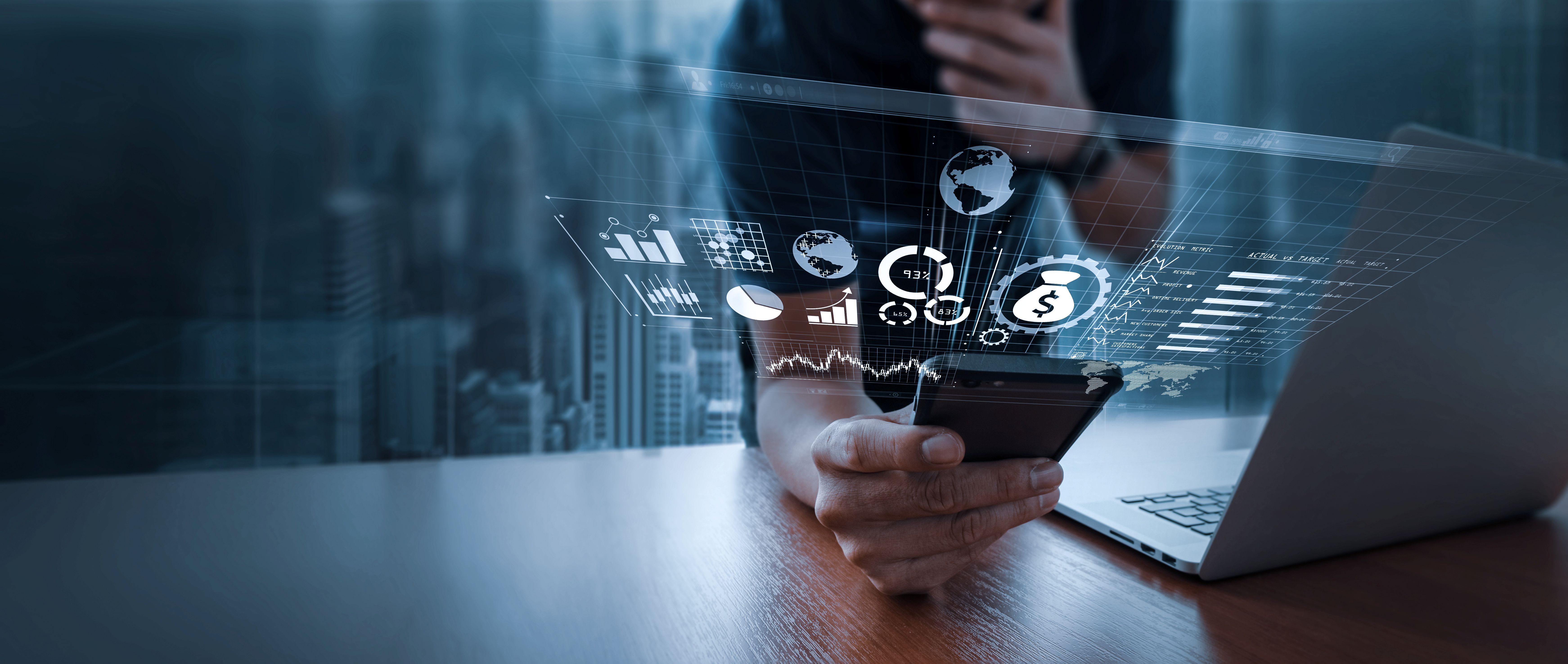 A person's hands hold a smartphone, from which a series of glowing digital infographics and data visualizations are projected into the air, including charts, globes, and a money bag icon. A laptop rests open on the table beside the phone, and a man's out-of-focus face is visible in the background, thinking. A blurred cityscape is seen through a window.