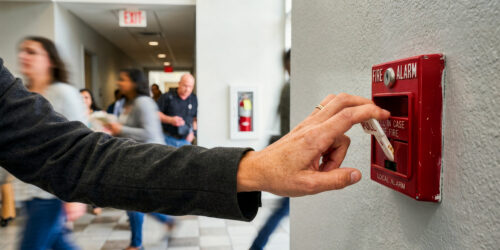 A close-up shot of a person's hand pulling down the white lever of a red fire alarm box mounted on a textured, light-colored wall. The alarm box has the words 'FIRE ALARM' and 'PULL IN CASE OF FIRE' printed on it. The person is wearing a dark grey long-sleeved shirt or jacket. In the blurred background, several people are walking down a brightly lit hallway, suggesting a public building, school, or office environment. An 'EXIT' sign and a fire extinguisher cabinet are also visible in the distance.