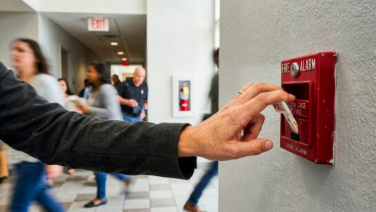 A close-up shot of a person's hand pulling down the white lever of a red fire alarm box mounted on a textured, light-colored wall. The alarm box has the words 'FIRE ALARM' and 'PULL IN CASE OF FIRE' printed on it. The person is wearing a dark grey long-sleeved shirt or jacket. In the blurred background, several people are walking down a brightly lit hallway, suggesting a public building, school, or office environment. An 'EXIT' sign and a fire extinguisher cabinet are also visible in the distance.