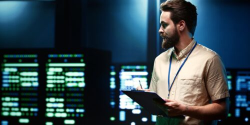 An IT specialist walking through a row of server racks in a data center, representing the infrastructure covered in a disaster recovery test
