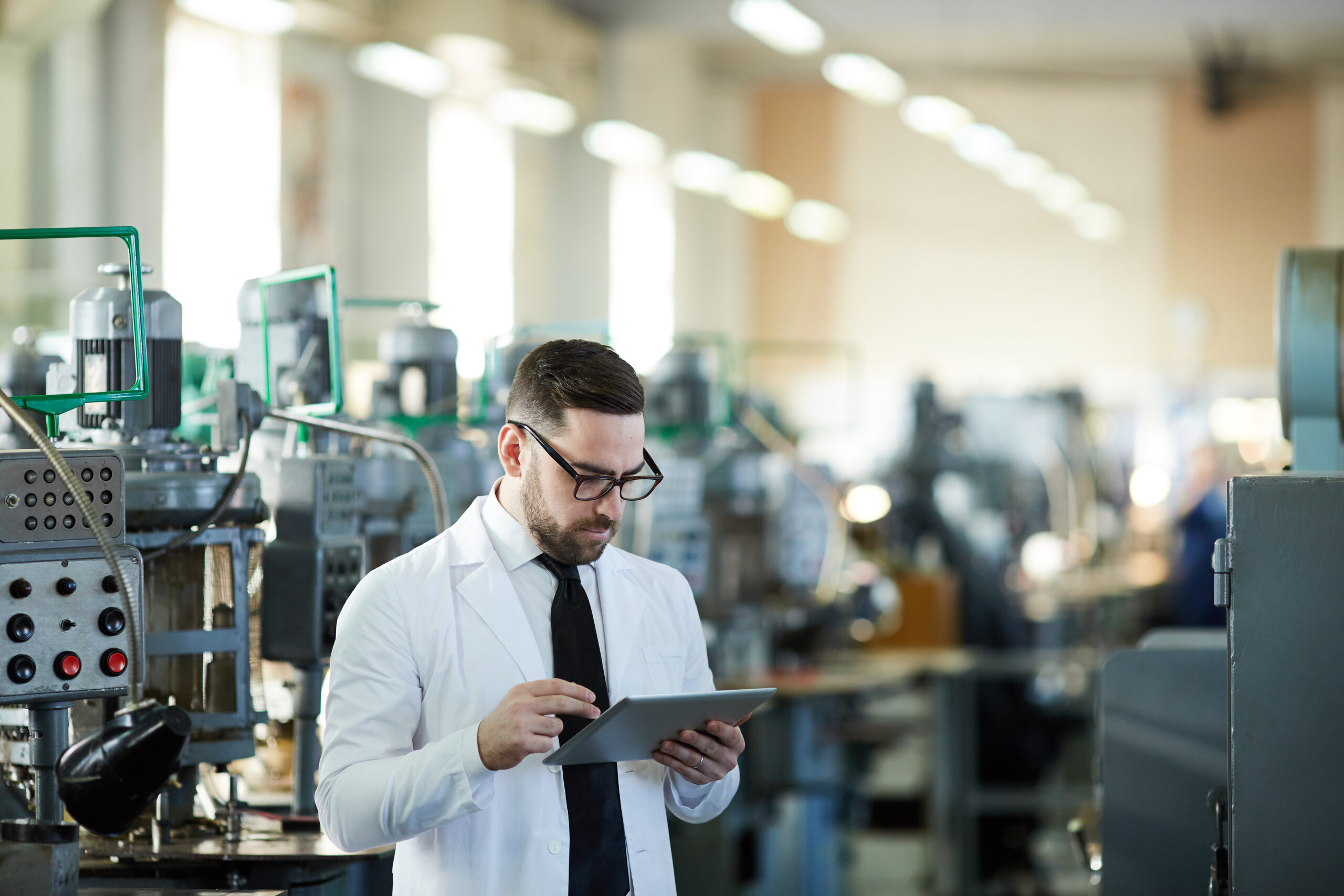 A factory worker in a lab coat reviewing information on a digital tablet in an industrial workshop
