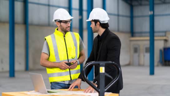Warehouse manager and staff member reviewing systems on a laptop in a distribution centre, the kind of operation where an in-house IT lead works with an outside team