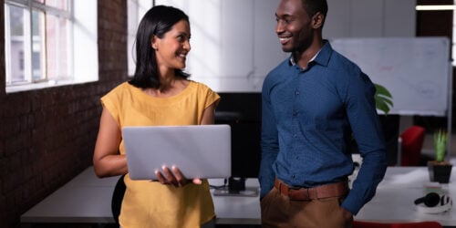 Two colleagues standing in an office looking at a laptop together, like an IT contact who already knows the team working through an issue with a staff member