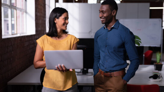 Two colleagues standing in an office looking at a laptop together, like an IT contact who already knows the team working through an issue with a staff member