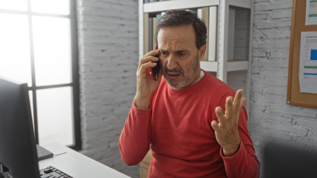 Frustrated business owner on the phone at his office desk, chasing his IT provider about a computer problem